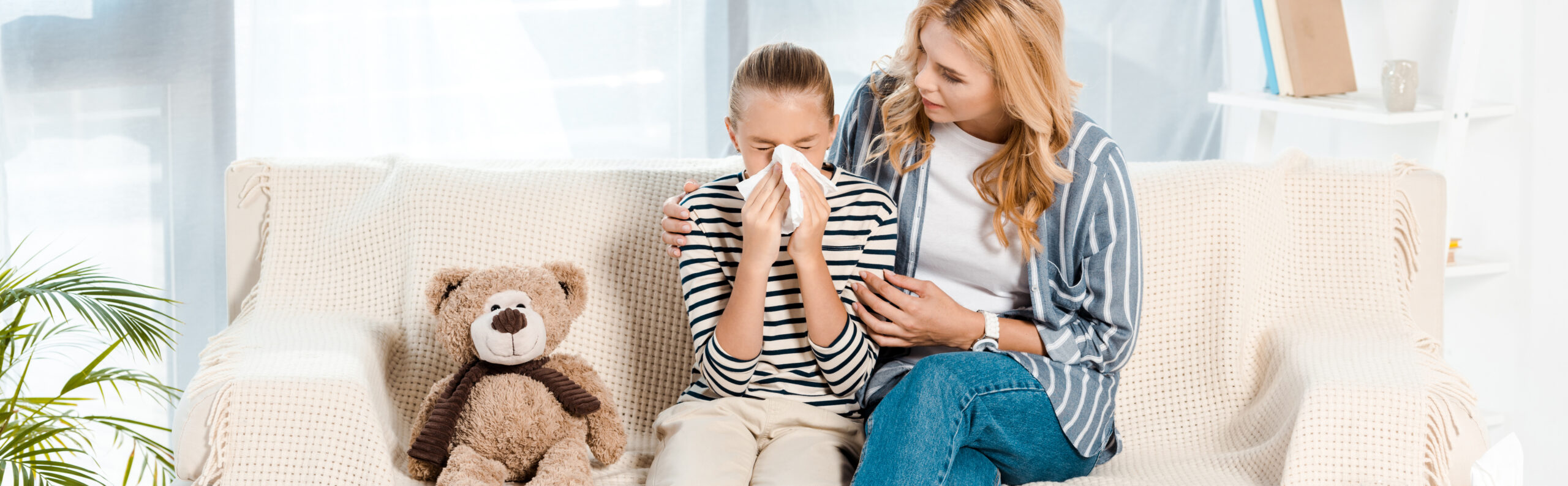 panoramic shot of woman sitting near daughter sneezing in tissue near teddy bear